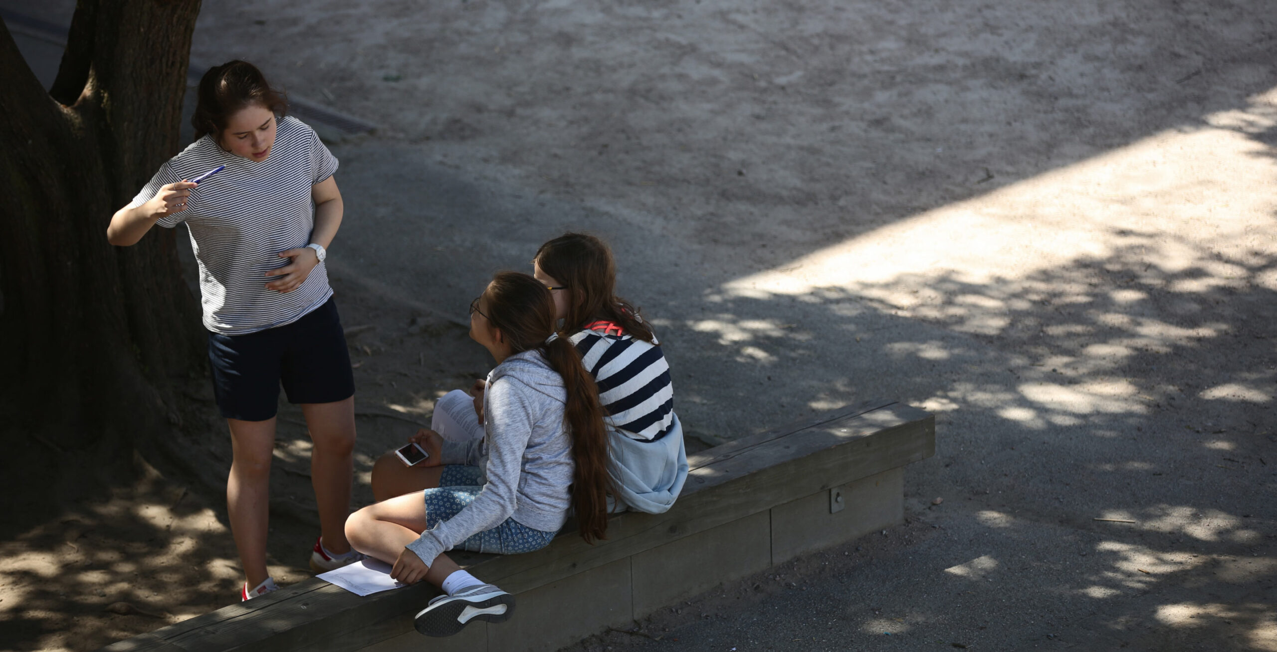3 Kinder unterhalten sich. Zwei sitzen auf einer Bank, das dritte steht vor ihnen und erzählt etwas.